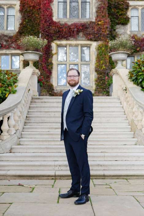 A bearded man in a navy suit with a boutonniere stands on stone steps in front of an ivy-covered building with large windows.