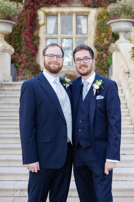Two men in navy suits with boutonnieres pose together on stone steps in front of an ivy-covered building.