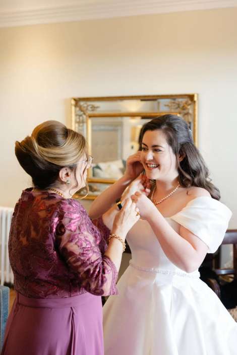 A bride in a white wedding dress smiles as an older woman in a purple lace dress helps her with jewelry.