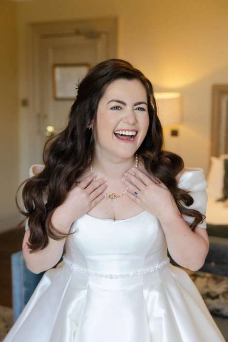 Joyful bride in a white wedding gown with a pearl necklace and beaded belt, laughing with her hands at her neckline.