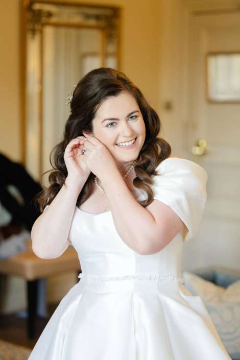 A bride in a white wedding dress smiles while putting on her earrings.