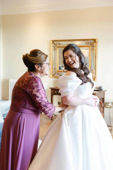 A bride in a white wedding gown smiles as an older woman in a purple lace dress helps adjust her dress in a warmly lit room.