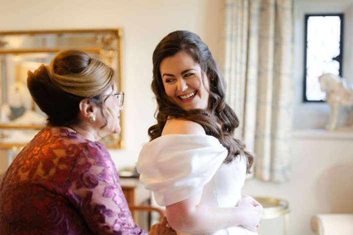 A smiling bride in a white dress hugs an older woman wearing a purple floral dress indoors.