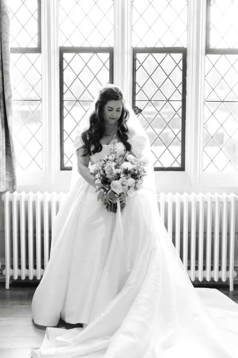 A bride in a flowing white wedding gown and veil holds a bouquet while standing in front of large diamond-pane windows and a radiator.