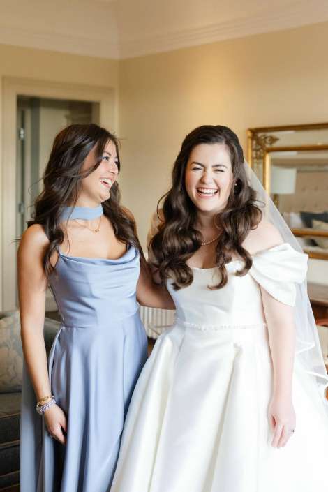 A bride in a white wedding gown with veil and a bridesmaid in a light blue dress share a joyful laugh indoors.