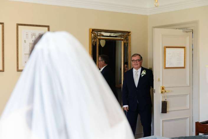A man in a dark suit with a white boutonniere stands in a doorway of a cream-colored room, smiling as a bride wearing a veil passes by in the foreground.