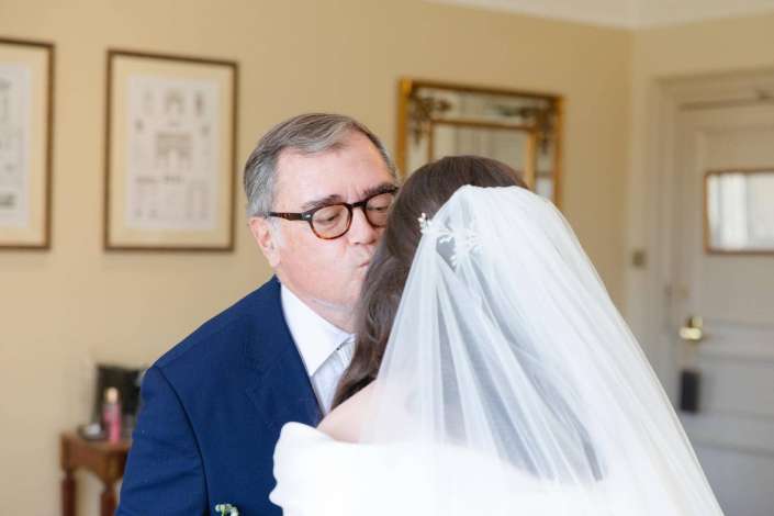 A groom in a dark suit gently kissing a bride in a white wedding gown and veil indoors.