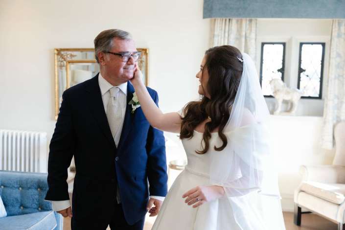Bride in a white wedding dress and veil gently touches the cheek of a smiling man in a navy suit indoors.