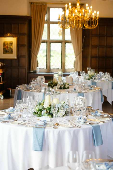 A formal dining room with round tables dressed in white and blue, glassware and candle centerpieces, illuminated by a chandelier.