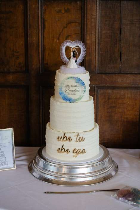 Three-tier white wedding cake with a heart-shaped topper and a blue wreath plaque reading MR. & MRS. Lansdale 25-10-2025, on a silver cake stand.