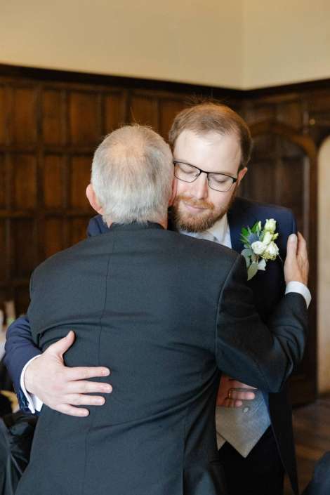 Two men in suits hug at a wedding, one wearing a boutonniere.
