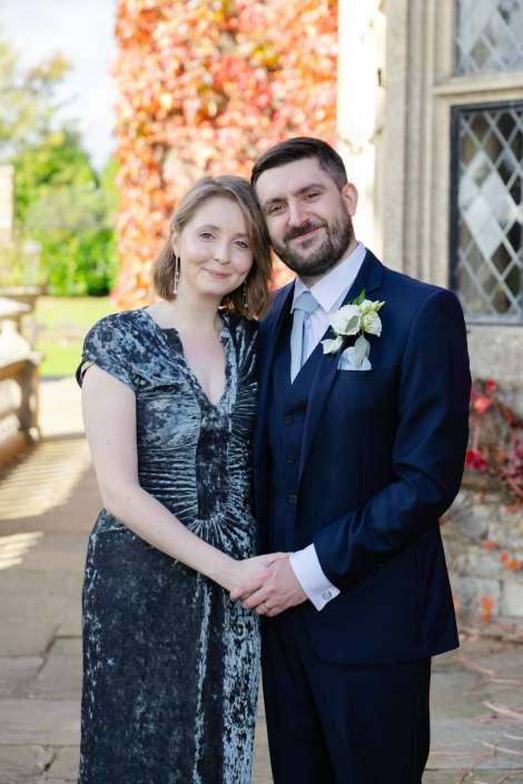 A smiling couple dressed in formal attire stands outdoors, the woman in a dark velvet dress and the man in a navy suit with a boutonniere, their hands clasped.