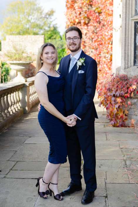 A smiling couple in navy formalwear stands outside, holding hands; the woman wears a navy dress and heels, the man a navy suit with a boutonniere.