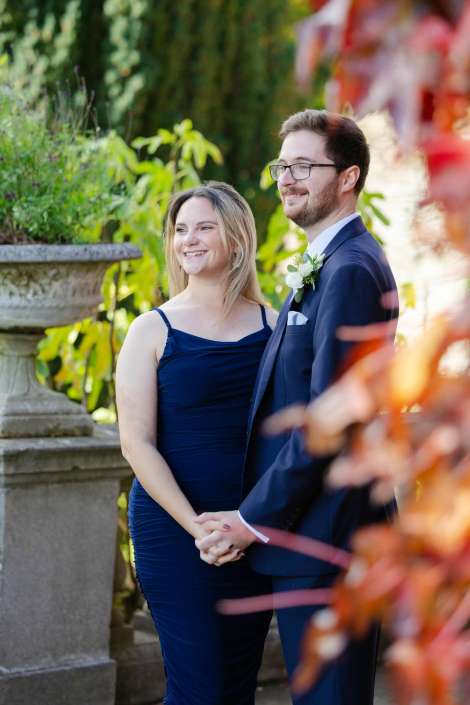 A smiling couple stands hand-in-hand in a garden, the woman in a navy dress and the man in a navy suit with a boutonniere.
