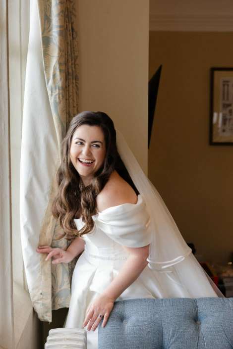 A joyful bride in an off-the-shoulder white wedding gown with a veil stands by a window, touching a patterned curtain.