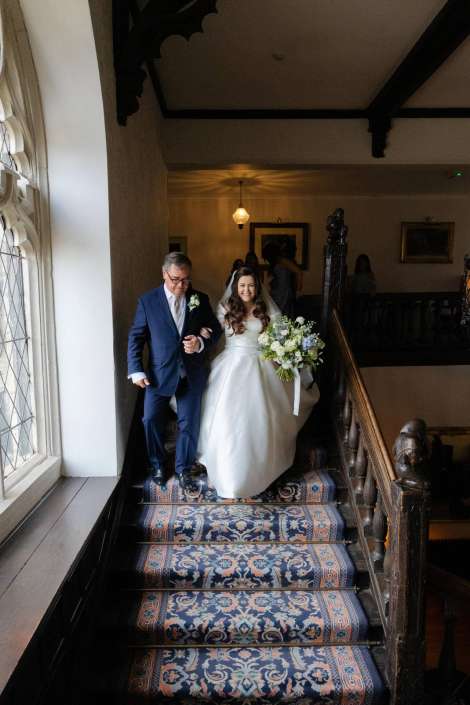 A bride in a white wedding gown carrying a bouquet descends a patterned carpeted staircase beside a man in a navy suit.