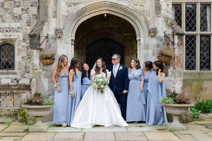 Bride and groom stand with bridesmaids in light blue dresses outside a historic stone building.