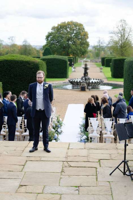 A man in a navy suit with a boutonniere stands at the head of a white aisle in an outdoor wedding ceremony, with guests seated and a fountain in the garden behind.