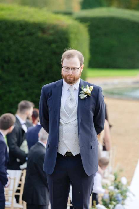 A man in a navy suit with a light gray vest and boutonniere stands with his hands behind his back at an outdoor wedding, with guests seated in the background.