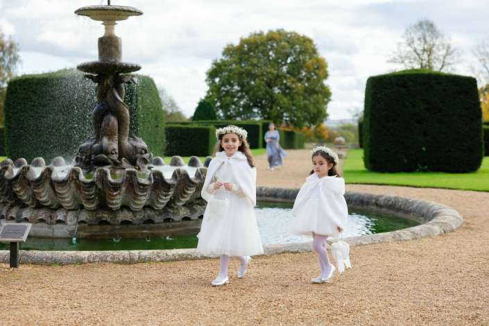 Two young girls in white dresses with flower crowns walk beside a large ornate fountain in a formal garden.