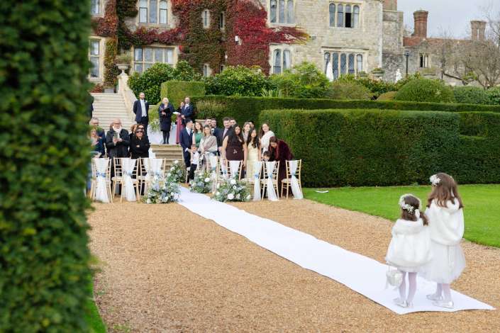 Wedding guests stand along a white aisle in a formal garden outside an ivy-covered stone manor.