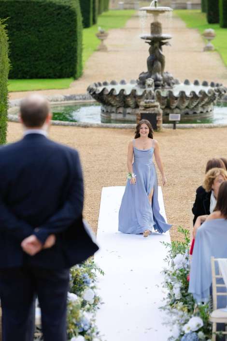 A smiling bridesmaid in a light blue gown walks down a white aisle runner at an outdoor garden wedding, with a fountain and seated guests in the background.