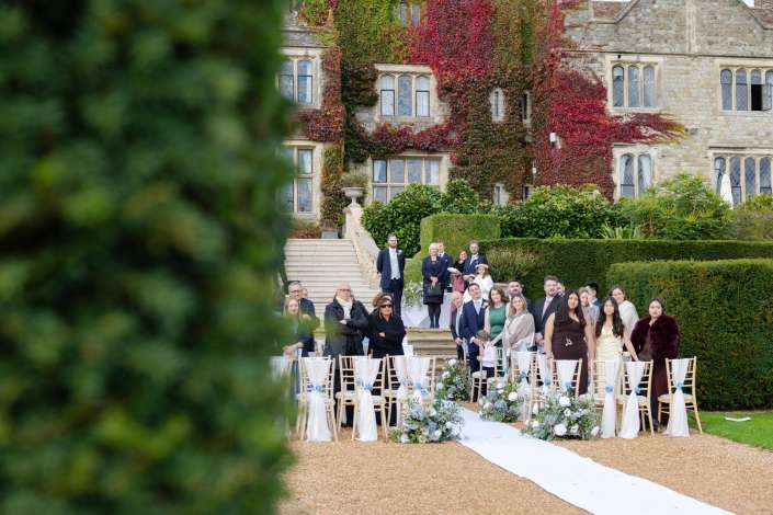 Wedding guests stand along a white aisle on a gravel lawn in front of an ivy-covered stone manor.