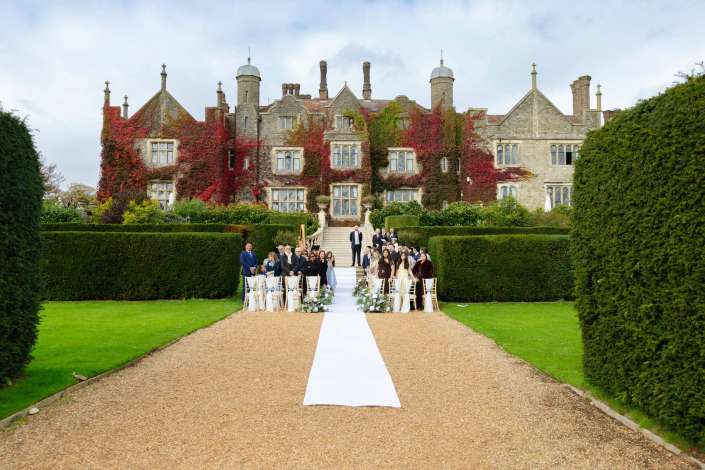 An outdoor wedding ceremony on a white aisle runner with guests gathered along the sides, in front of a large ivy-covered stone mansion.