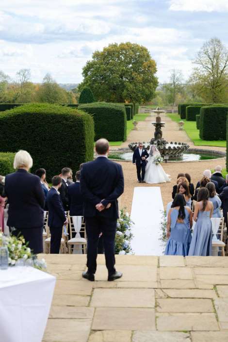 Bride and groom walk down a white aisle runner toward a fountain in a formal garden, as guests sit on either side.