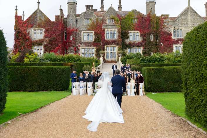 Bride and groom walk down a gravel wedding aisle toward their guests outside an ivy-covered stone mansion.