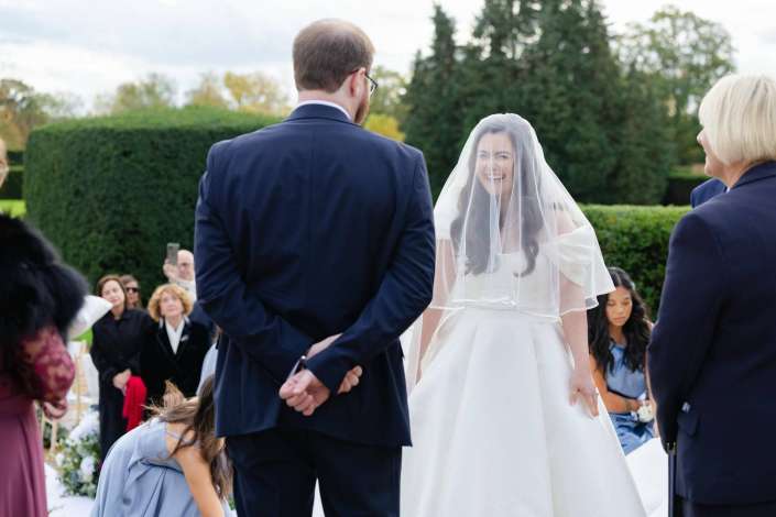 Bride in a white wedding gown and veil smiles at her groom in a dark suit outdoors during a wedding ceremony.