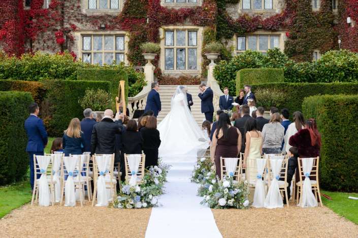 Outdoor wedding ceremony on a white aisle with floral arrangements, in front of an ivy-covered stone mansion, with the bride and groom at the steps and guests gathered.