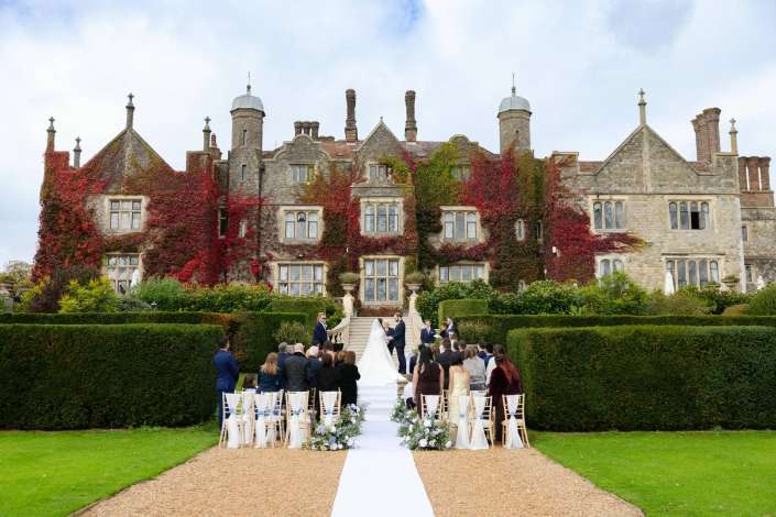 An outdoor wedding ceremony on a white aisle in front of an ivy-covered historic stone mansion, with guests gathered.
