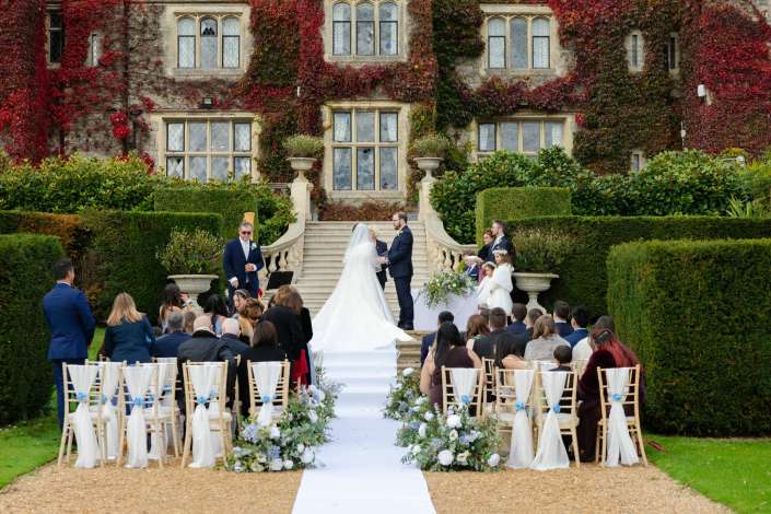 An outdoor wedding ceremony on a gravel aisle before an ivy-covered historic stone building, with the bride and groom at the altar as guests look on.