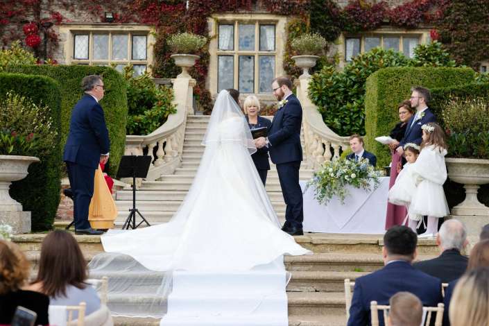 Bride and groom exchange vows with an officiant on a grand stone staircase at an outdoor wedding, surrounded by guests and lush greenery.