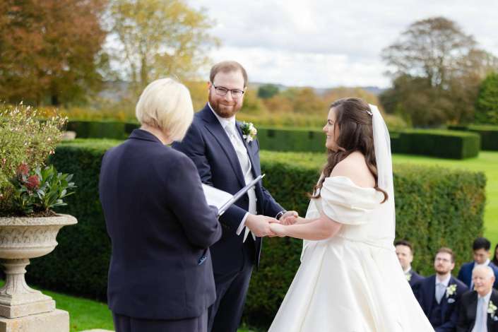 Bride in a white wedding dress and veil and groom in a dark suit exchange rings outdoors with an officiant overseeing.