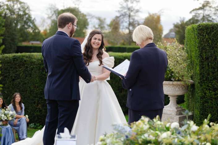 Smiling bride in a white wedding gown shakes hands with her groom outdoors, while an officiant holds a binder and bridesmaids in blue dresses sit nearby.