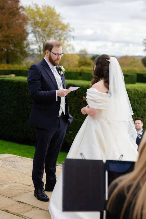 Groom in a navy suit reads from a card to a bride in a white off-the-shoulder gown with a veil, outdoors on a stone patio.