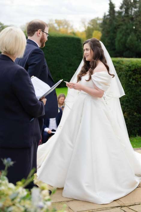 Bride in a white wedding gown with a veil stands with her groom in a dark suit during an outdoor wedding ceremony.