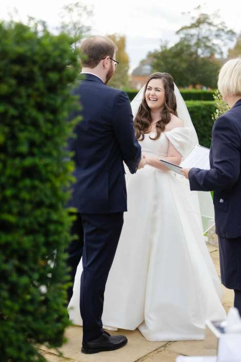 Bride in a white wedding gown and veil smiles while shaking hands with a groom in a dark suit at an outdoor garden ceremony.