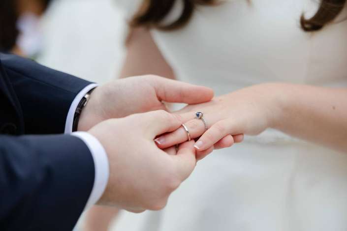 A close-up of a couple holding hands, with a silver ring featuring a blue gemstone on the woman's finger.