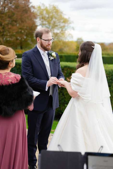 A groom in a navy suit places a ring on the bride's finger during an outdoor wedding ceremony.