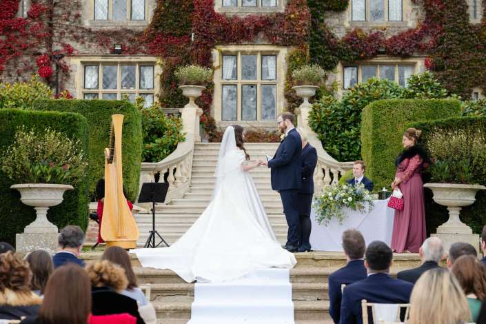 Bride and groom exchange vows on a grand stone staircase in front of an ivy-covered historic building, with bridesmaids, a harpist, and seated guests nearby.
