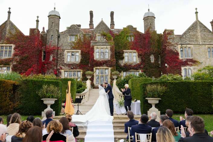 Bride and groom share a kiss during an outdoor wedding ceremony on a white aisle in front of an ivy-covered stone mansion with guests seated.