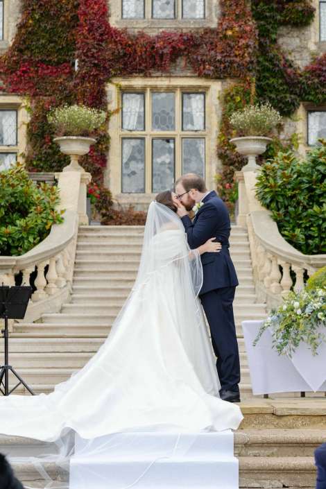 Bride and groom kiss on a grand stone staircase in front of an ivy-covered historic building, with the bride in a white wedding gown and veil.