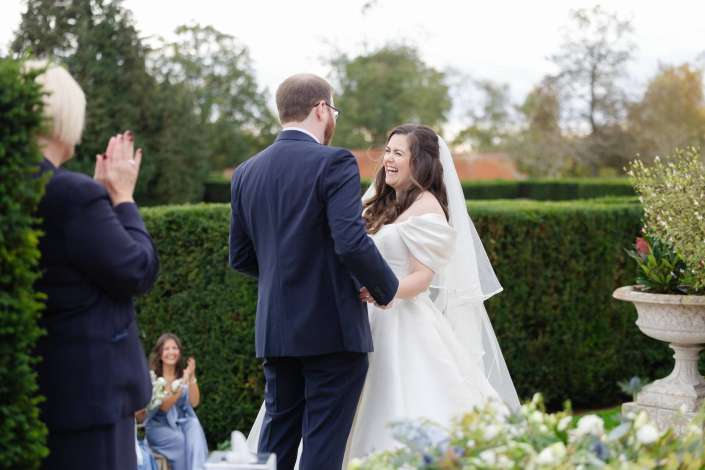 Bride in a white wedding gown and veil and groom in a navy suit share a joyful moment outdoors as guests applaud.