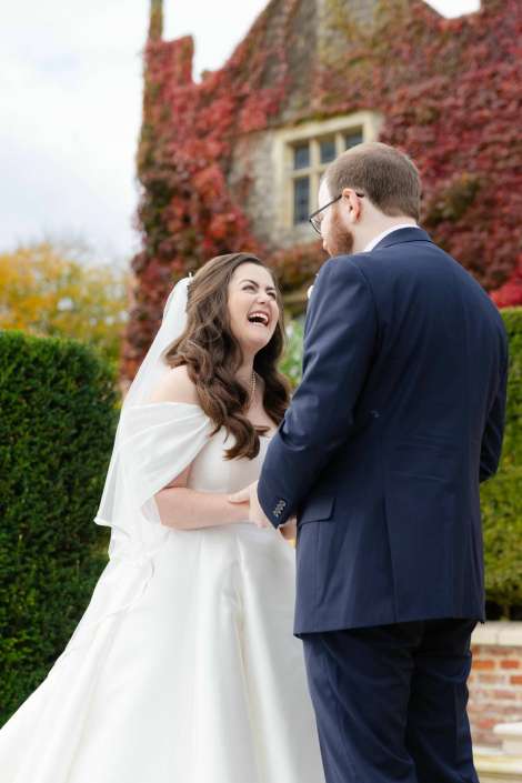 Bride in a white wedding dress and veil laughs while holding hands with a groom in a navy suit outdoors, with an ivy-covered building in the background.
