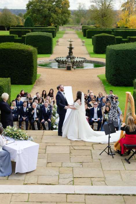 Bride and groom exchange vows during an outdoor garden wedding, with a central fountain and neatly trimmed hedges as guests look on.