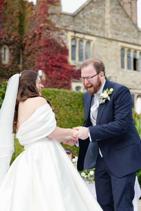 Joyful bride in a white wedding gown and veil and groom in a navy suit hold hands and laugh at their outdoor wedding in front of a stone building.