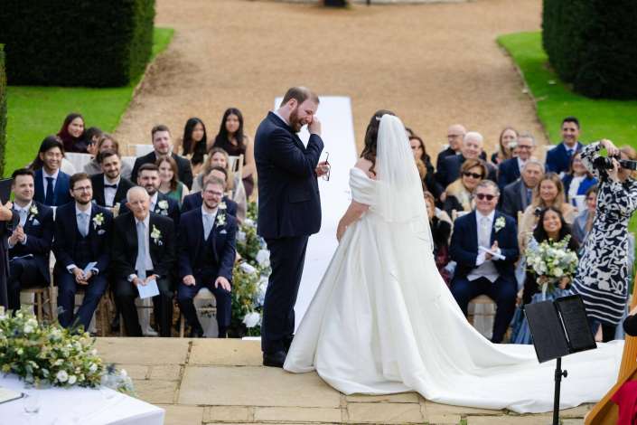 Groom wiping a tear as the bride in a white wedding gown and veil stands before him at an outdoor ceremony with guests seated along the aisle.